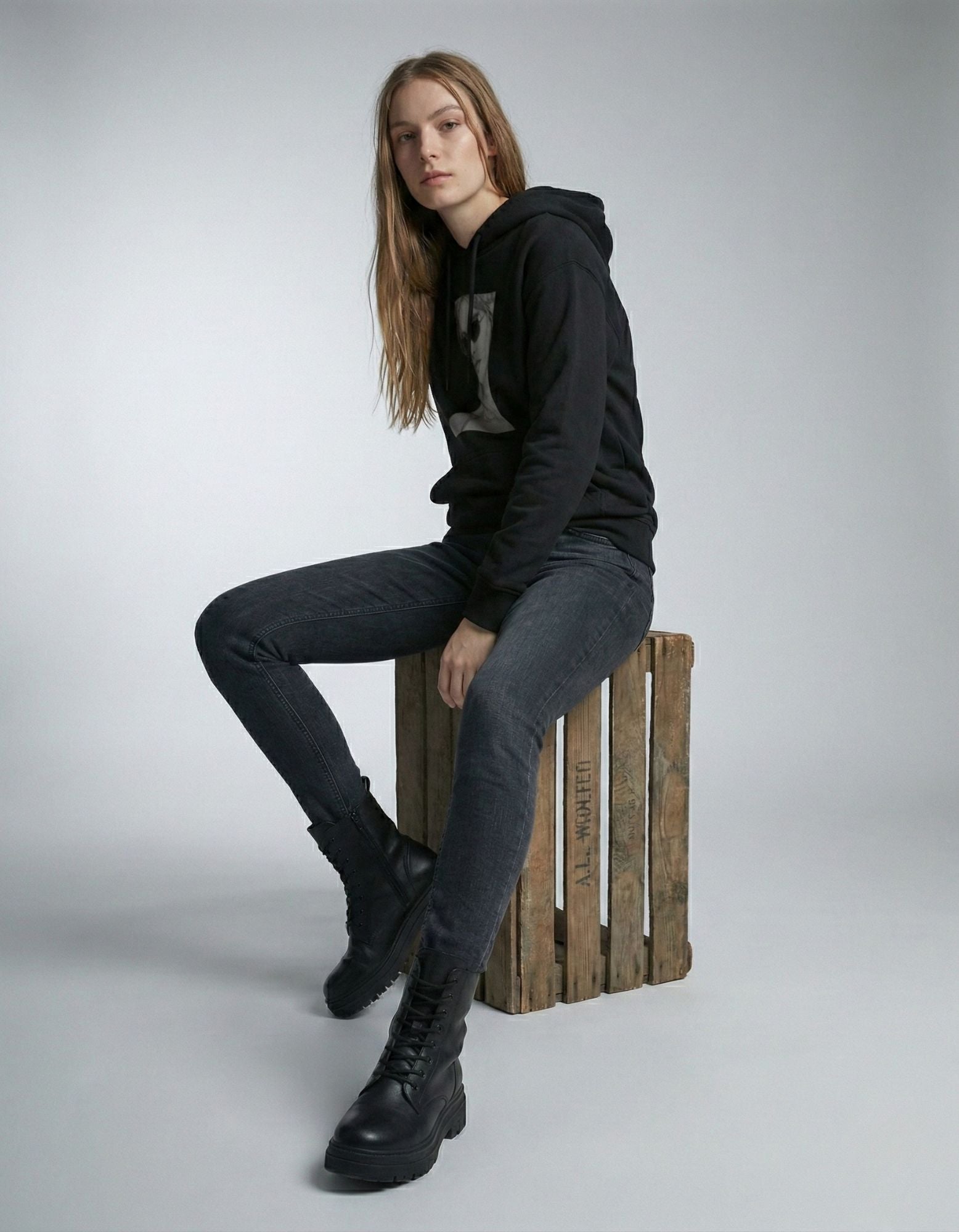 Female model sitting on a wooden crate wearing a black GENZs Fashion graphic hoodie and dark slim-fit jeans.