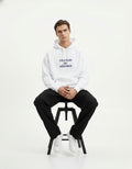 A male model sitting on a stool wearing a GENZs Fashion white hoodie with "STREETWEAR, NOT SWEETWEAR" printed on the chest.