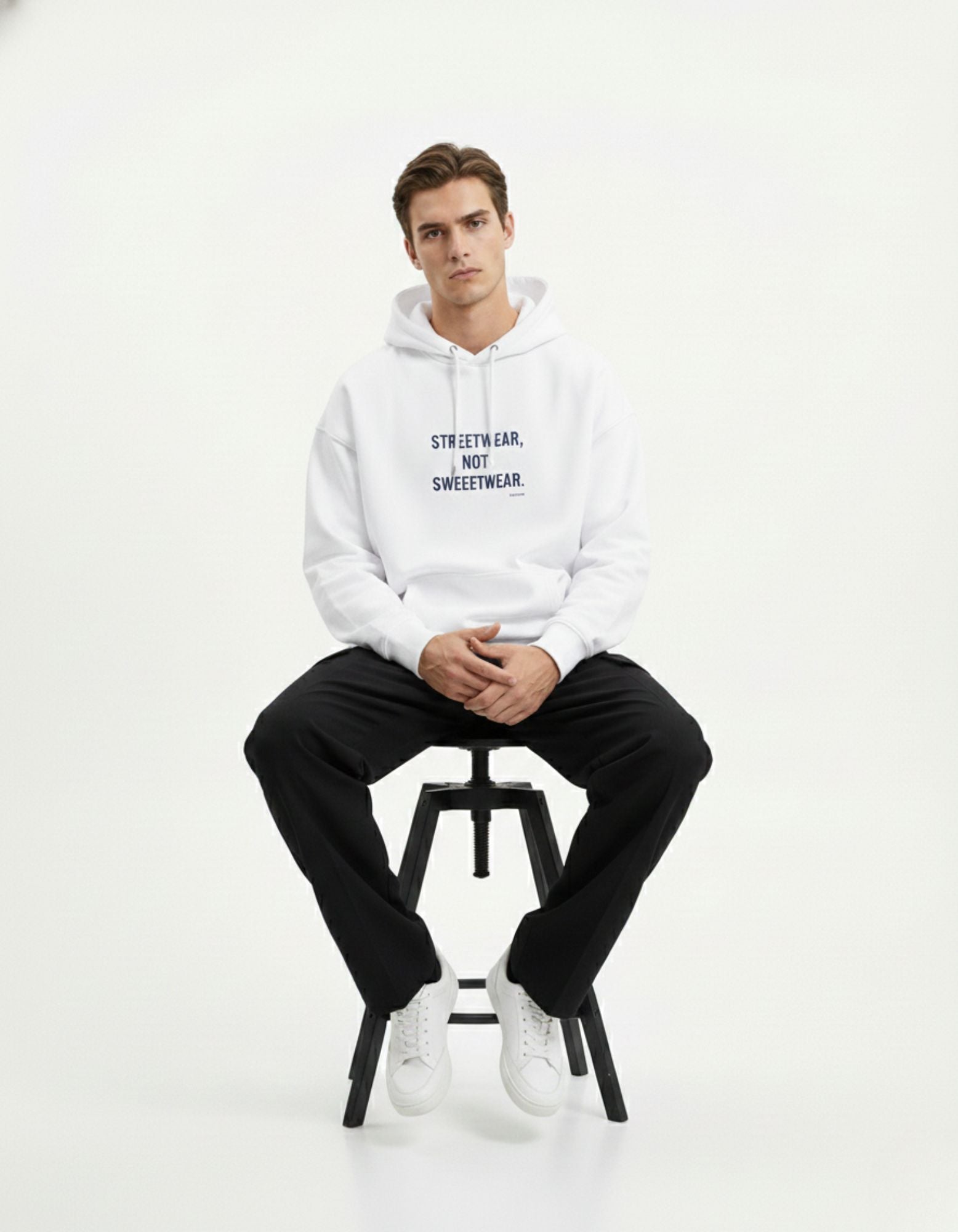 A male model sitting on a stool wearing a GENZs Fashion white hoodie with "STREETWEAR, NOT SWEETWEAR" printed on the chest.