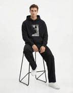 Male model sitting on a stool wearing a black heavy-weight graphic hoodie by GENZs Fashion.
