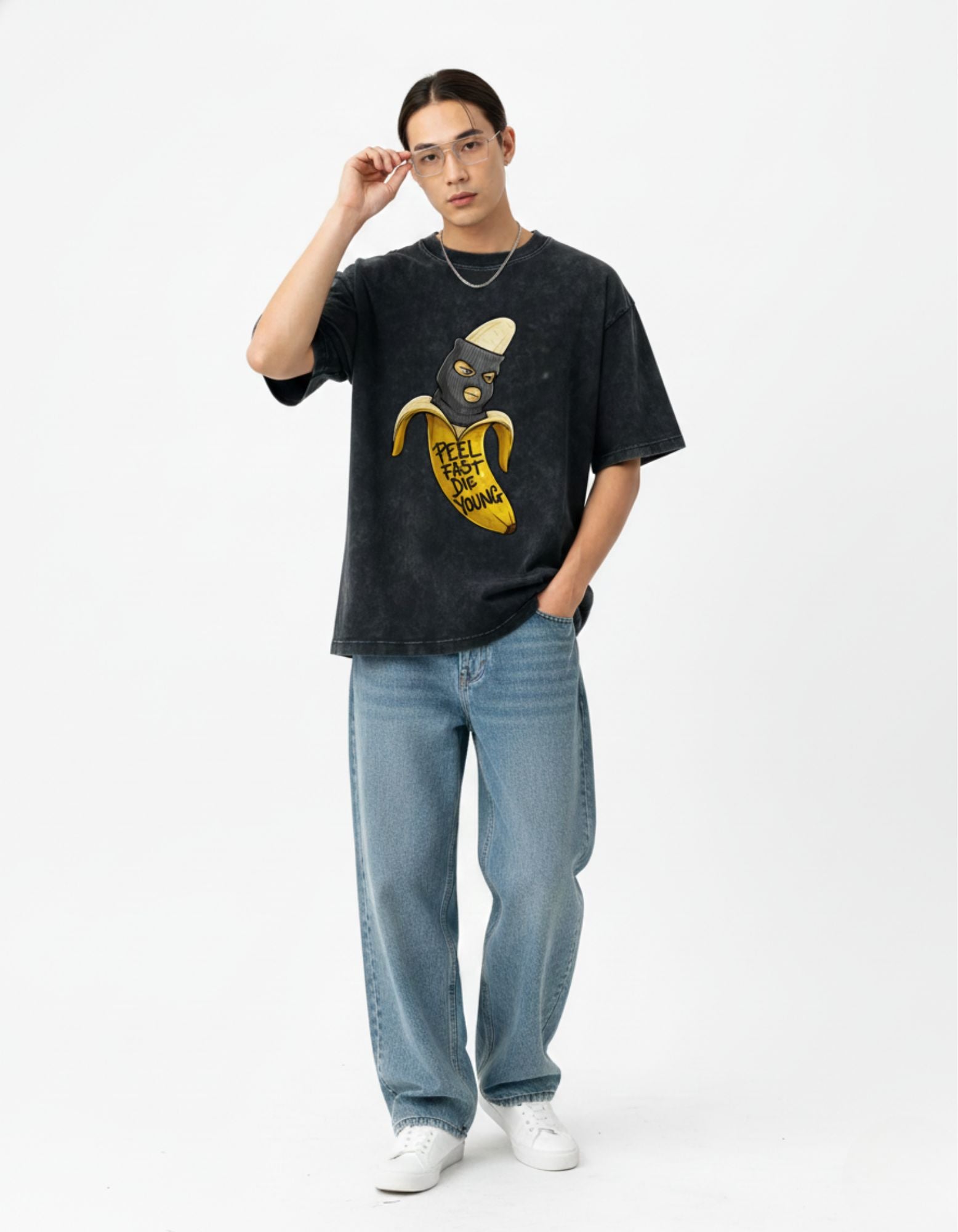 Man wearing a charcoal oversized t-shirt with a 'Peel Fast Die Young' banana mask graphic and baggy jeans.