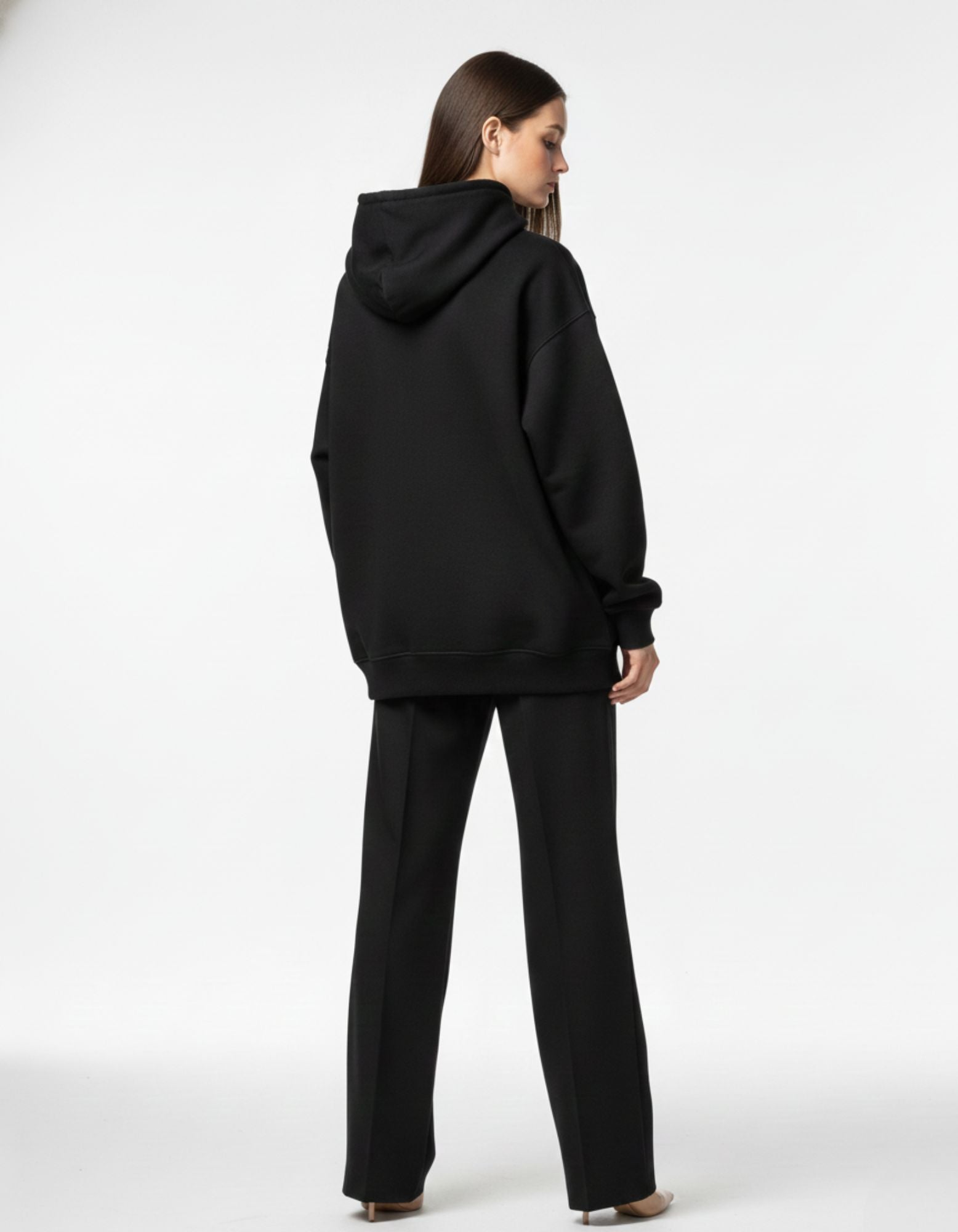 Rear view of a baggy black unisex hoodie worn with black trousers, highlighting the oversized streetwear fit.