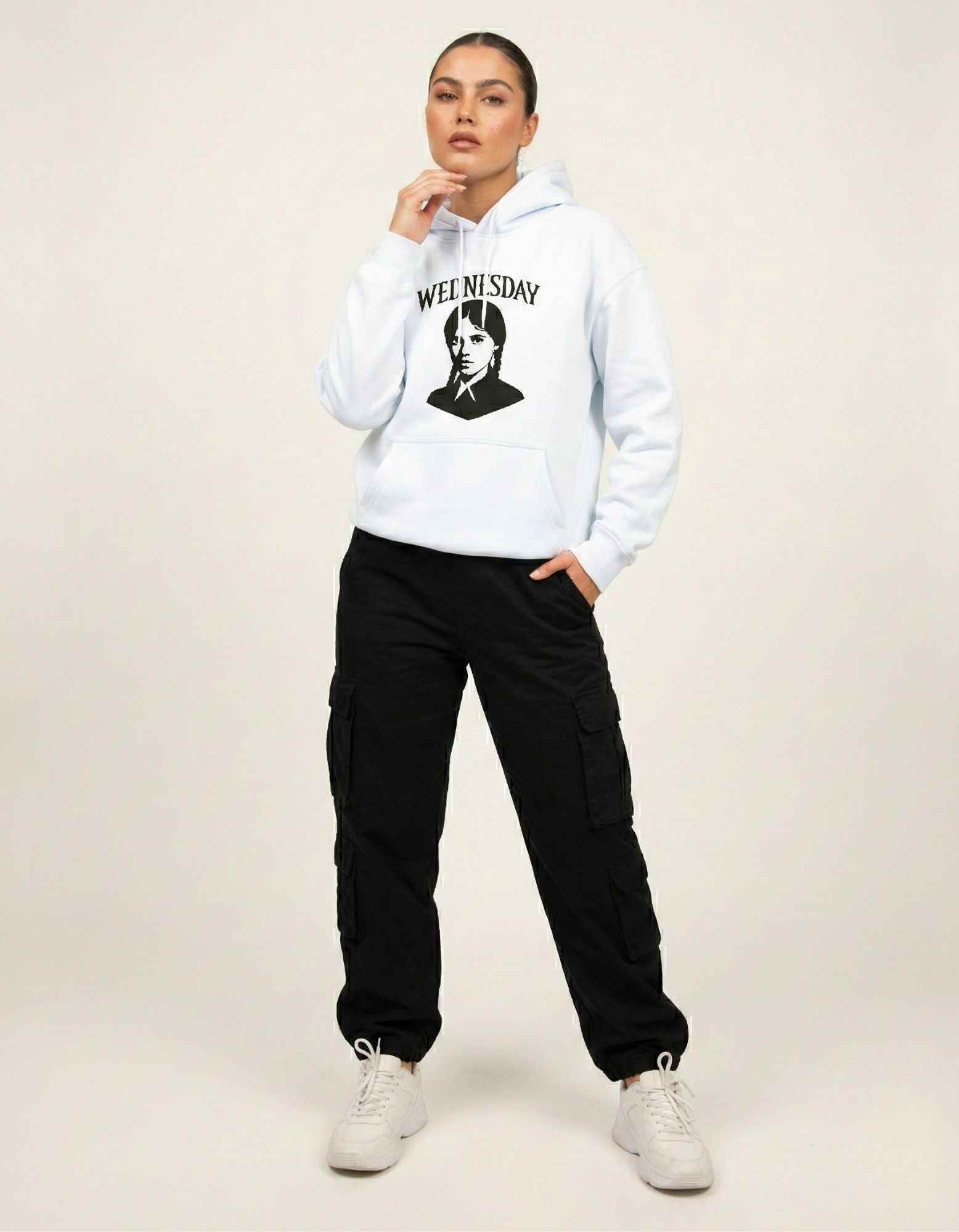 Female model standing in a light blue Wednesday Addams graphic hoodie with black cargo pants and white sneakers.