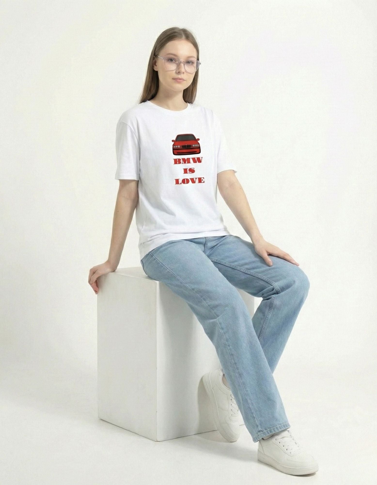A female model sitting and posing in the white 'BMW IS LOVE' graphic t-shirt, perfect for car culture and aesthetic streetwear.