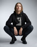 Female model squatting in a GENZs Fashion black hoodie featuring a grayscale portrait graphic print.