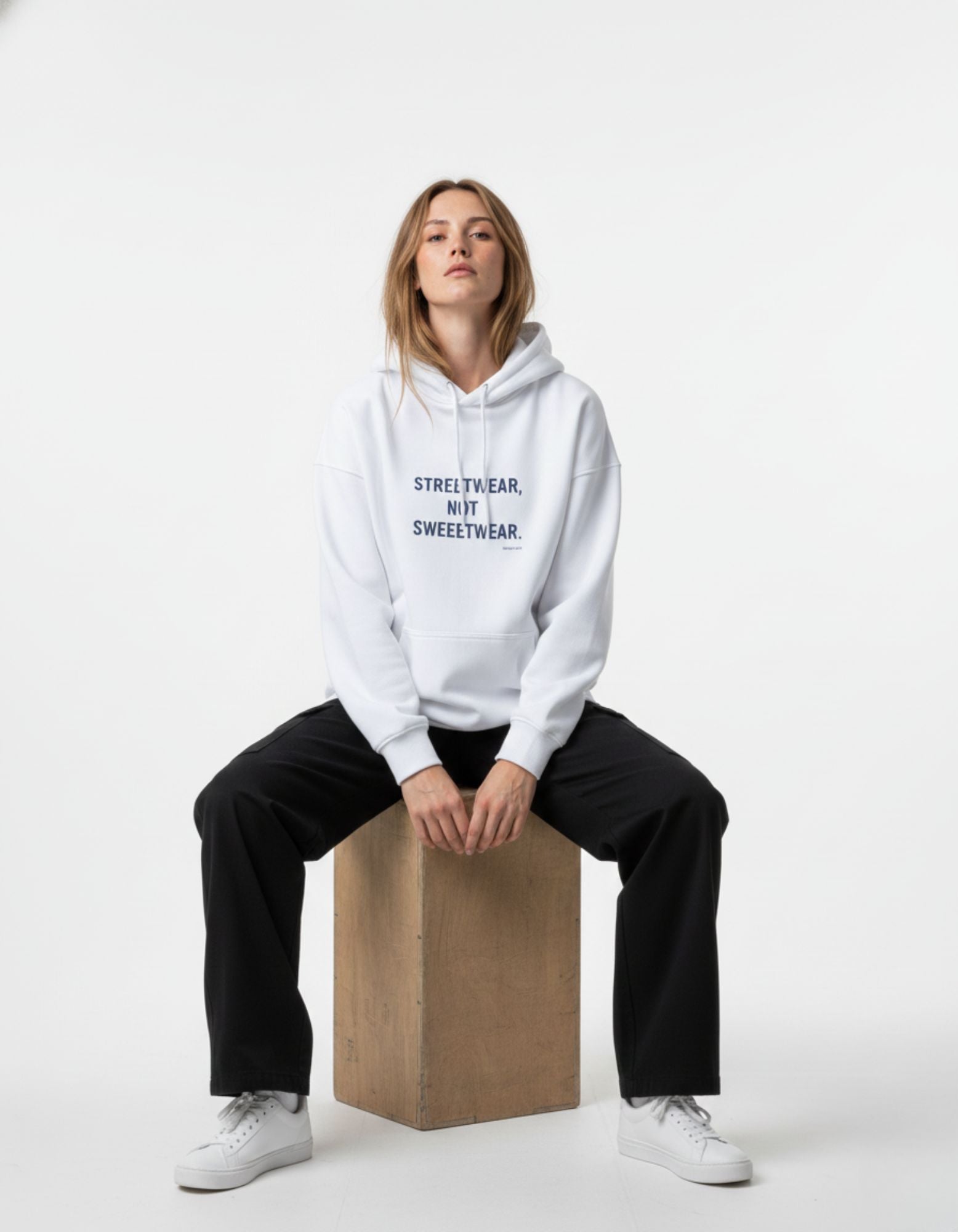 A female model sitting on a wooden box wearing an oversized white streetwear hoodie with a minimalist text logo.