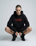 Female model in a crouching pose wearing a black Stranger Things branded hoodie with a red logo print.
